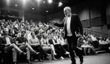 Eamon Blackthorn pacing the stage with a microphone before a packed tiered auditorium audience